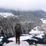 man with backpack on top of cliff staring at mountain